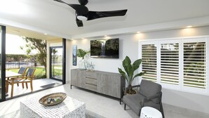 A modern living room with a grey sofa, a patterned ottoman, a wooden coffee table, and a grey cabinet. There is a large window with white blinds, a ceiling fan, and a TV mounted on the wall.