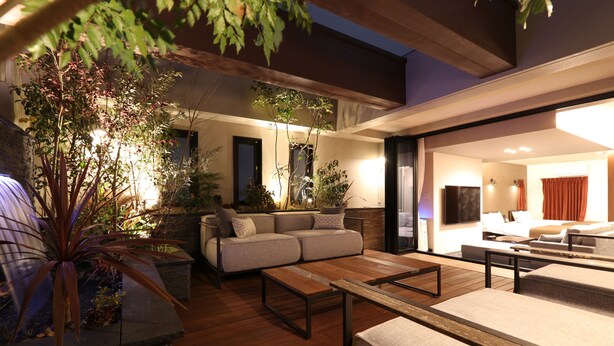 A modern living room with a sofa, coffee table, and plants. There is a dining area with a table and chairs. The room has a large window and a door leading to another room. - Amistad Hotel Fukuoka (Fukuoka)