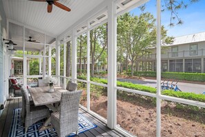 Un espacio exterior techado con una mesa y sillas, ventanas amplias y vistas a la calle y árboles.
