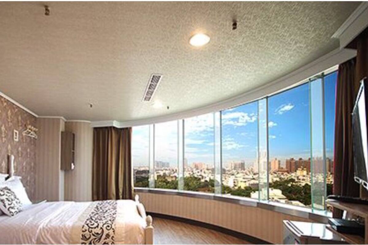 A hotel room with a large bed, a television, and a city view through the window.