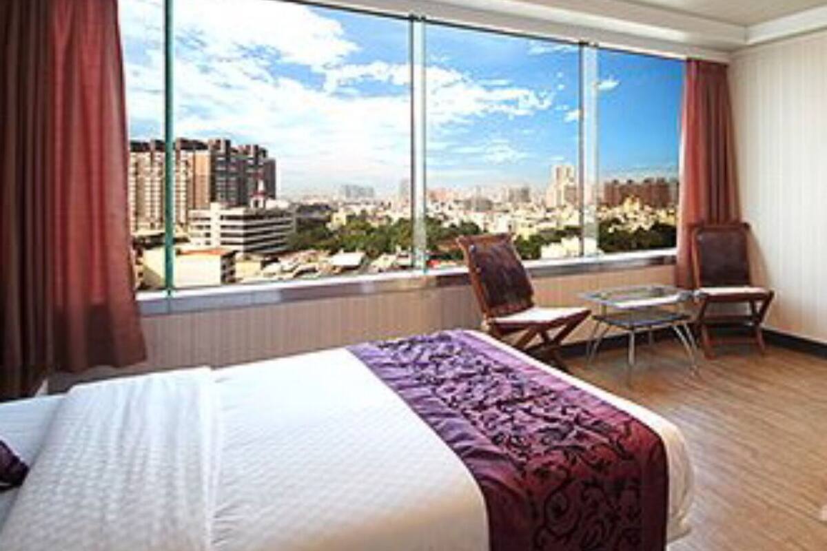 A hotel room with a large bed, a chair, a small table, and a view of a cityscape through large windows.
