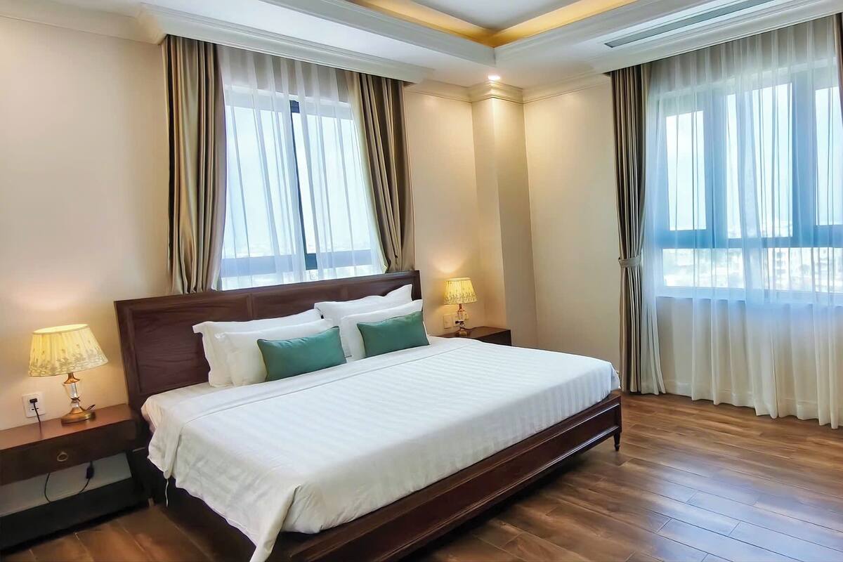 Une chambre d’hôtel avec un grand lit, deux lampes de chevet, une tête de lit en bois et une vue dégagée à travers des rideaux légers.