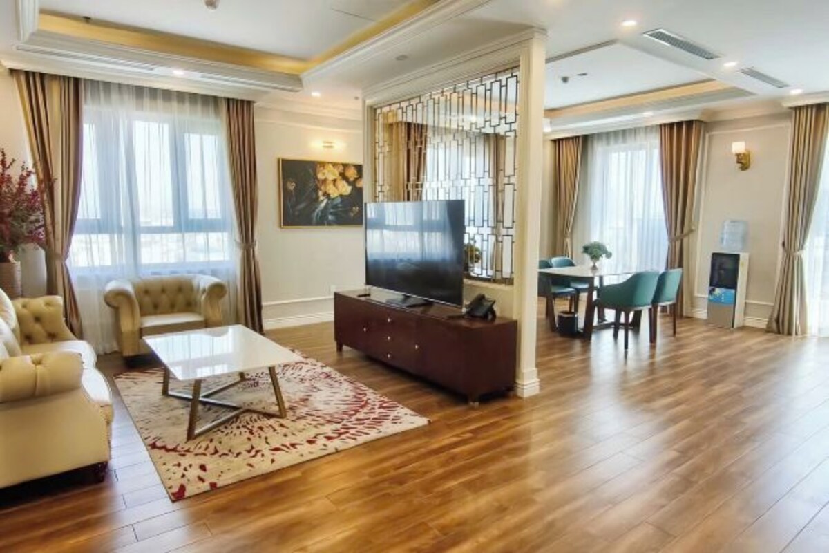 A spacious living room with a wooden floor, a sofa, a coffee table, a TV on a cabinet, and a dining area with chairs.