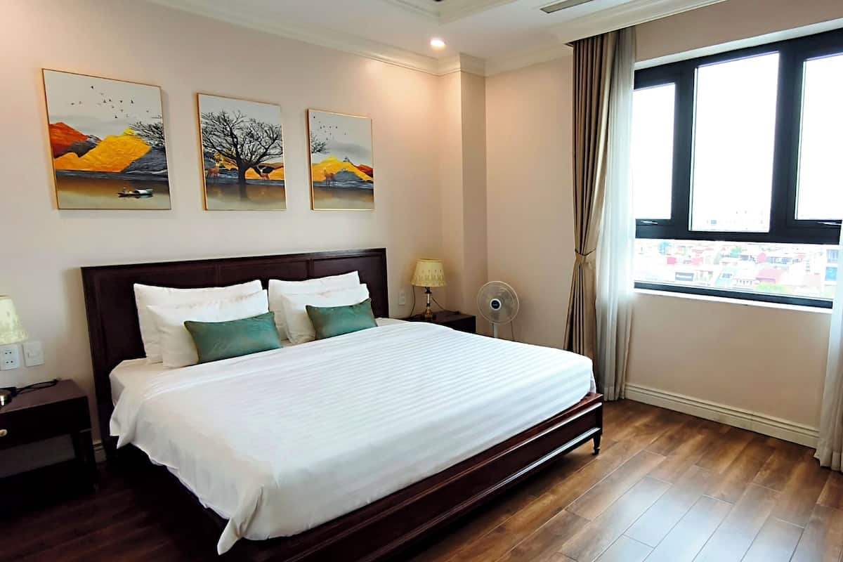 Une chambre d’hôtel avec un grand lit, une table de chevet avec une lampe, un ventilateur fixé au mur et une fenêtre avec des rideaux.