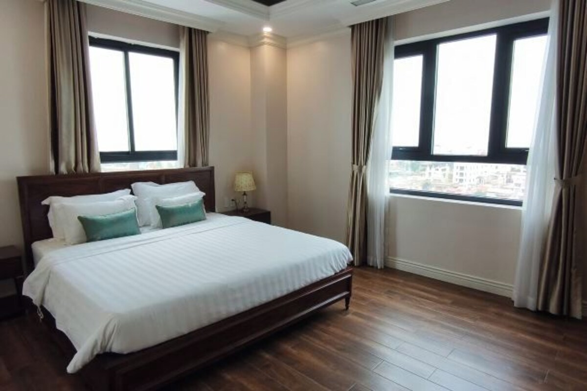 A hotel room with a large bed, two windows, a nightstand, and a view of the city.