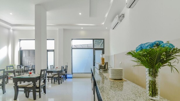A modern dining area with tables and chairs, a kitchen counter with a vase of flowers, and a large window providing natural light. - Royal Residence Golf View (Gurugram)