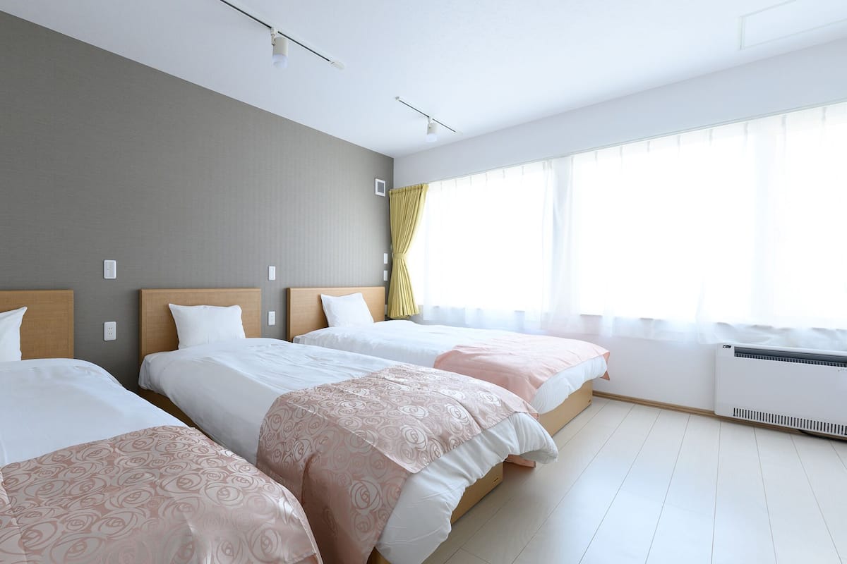 Ein Hotelzimmer mit drei Einzelbetten, einem großen Fenster mit Vorhängen, einer Klimaanlage und einem Lichtschalter an der Wand.