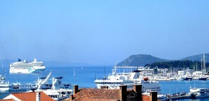 Ein Yachthafen mit mehreren angesterten Booten, ein Kreuzfahrtschiff im Hintergrund und eine gebirgige Landschaft.