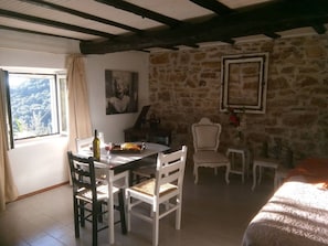 A room with a stone wall, a bed, a dining table with chairs, a small table with a vase, and a painting on the wall. - "Aristidis Stone House" (Gimari)