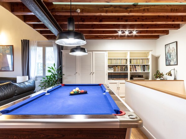 Een pooltafel met een blauwe viltkleed, omringd door houten kasten en een boekenplank vol boeken.
