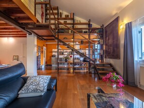 Een ruime woonkamer met een houten trap, een zwarte leren bank, een glazen salontafel en een keuken op de achtergrond.