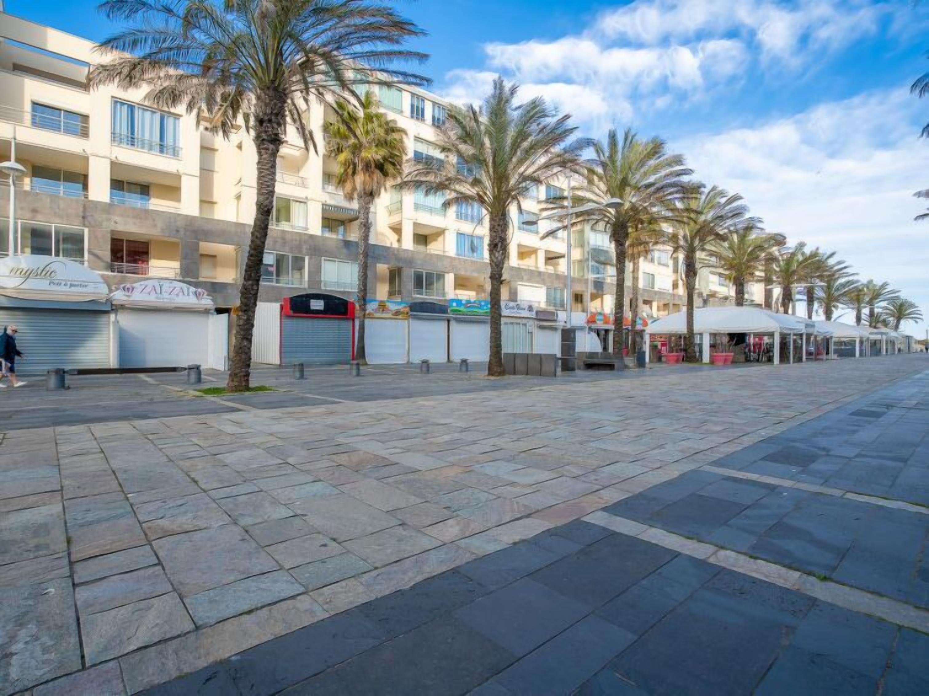 A beachfront promenade with palm trees, a row of closed shops, and a building with balconies.