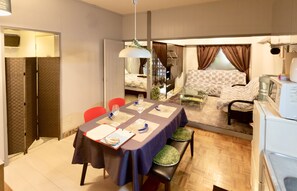 A compact room with a dining table set for four, a sofa, a piano, and a small kitchenette. - Holiday Homes TSUBOYA 6th (Naha)