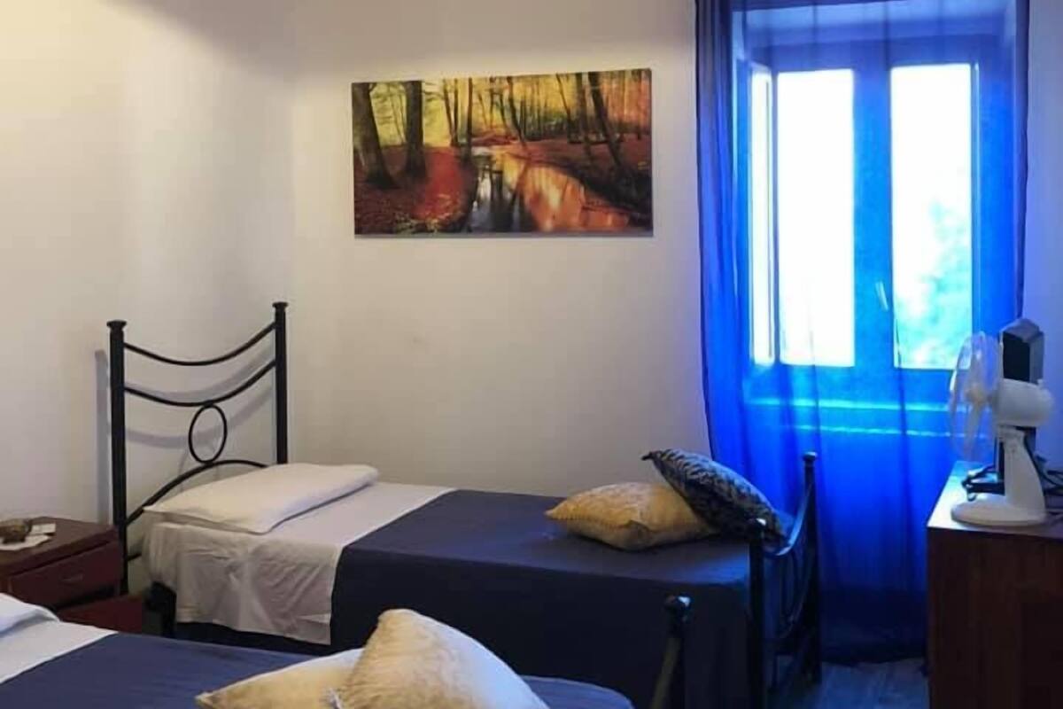 Una habitación con dos camas, un cuadro en la pared, una ventana con cortinas azules y un escritorio con una lámpara.