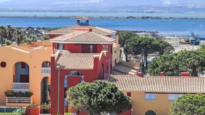 Un edificio costero con fachada roja y tejados de tejas de terracota, con vista a una masa de agua y montañas distantes.