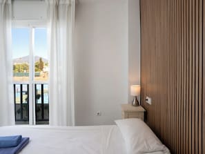 A hotel room with a bed, a nightstand, a lamp, a window with curtains, and a view of the outdoors. - Lorcrimar by Interhome (Marbella)