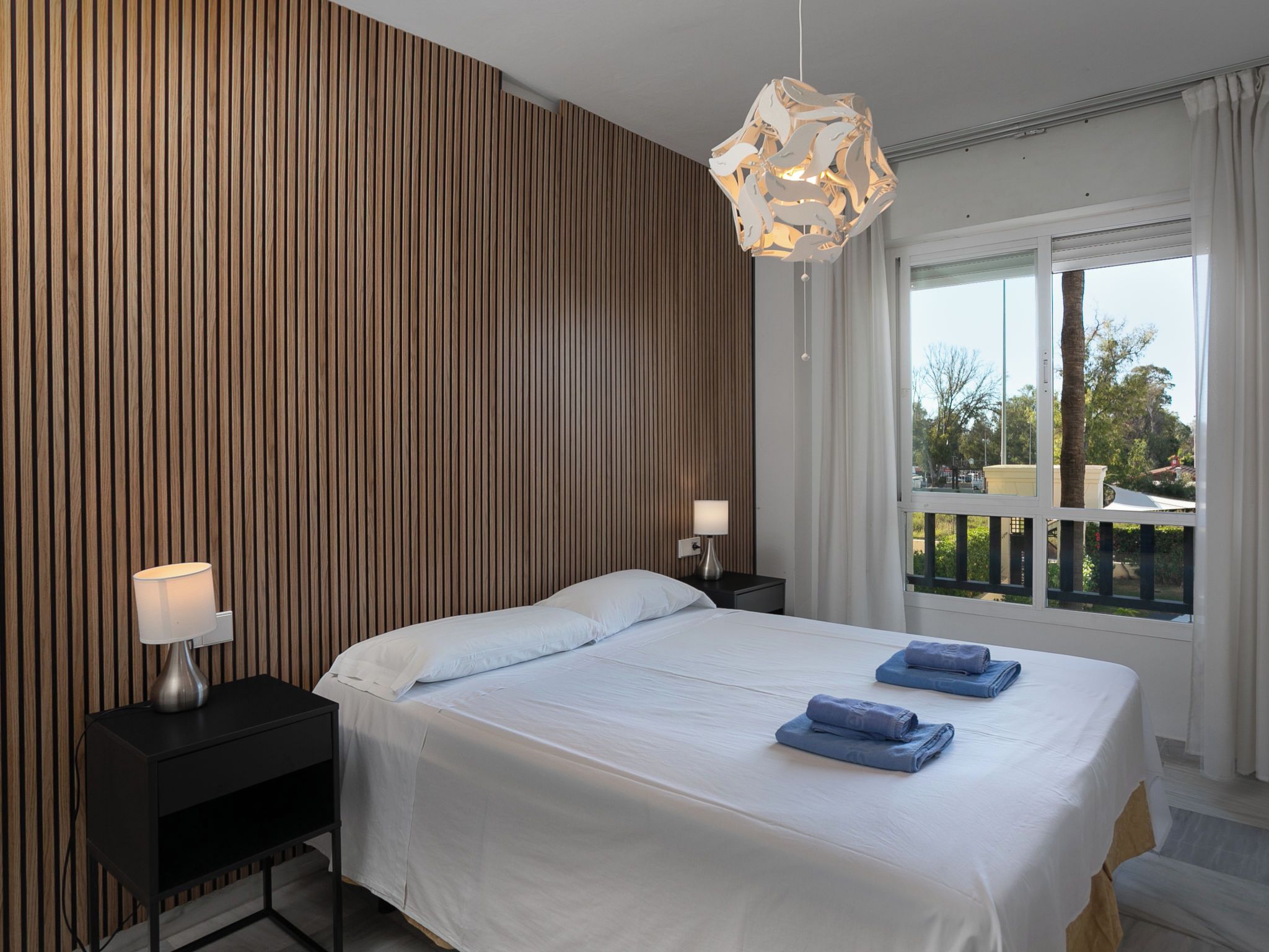 Een hotelkamer met een bed, twee nachtkastjes, een moderne hanglamp en een raam met uitzicht op het omliggende landschap.