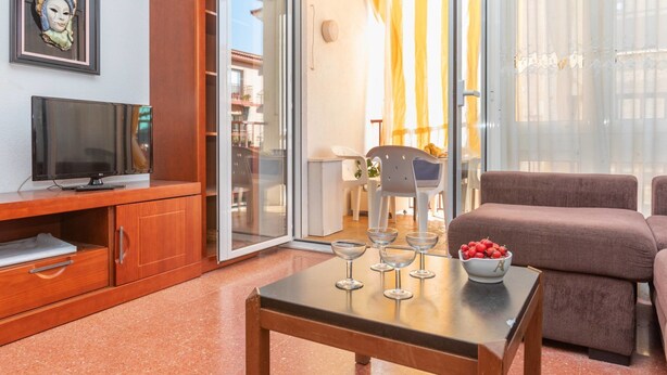 Una sala de estar moderna con televisor, muebles de madera, un ventanal de vidrio que da a un balcón y un área de comedor con cortinas amarillas.