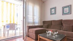 Una sala de estar con un sofá marrón, una mesa de centro de vidrio con copas de vino, y vistas a un espacio exterior con cortinas amarillas a través de un ventanal.