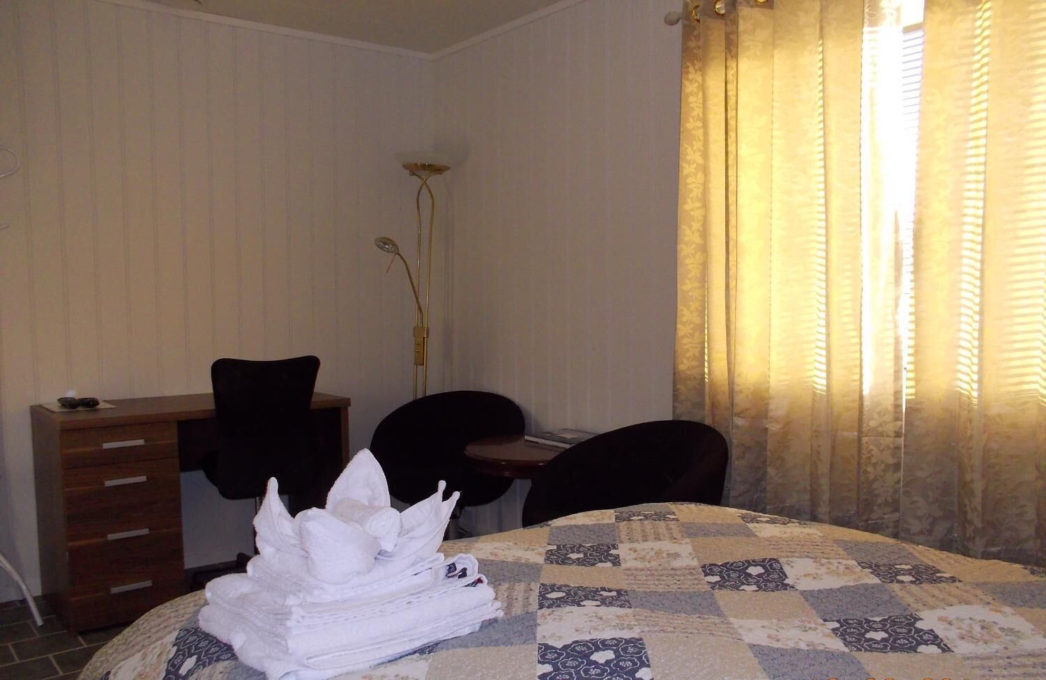 A hotel room with a bed, a desk, two chairs, a tall floor lamp, and a window with curtains.