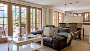 A spacious living area with a leather sofa, a glass coffee table, and a dining table with wicker chairs. Large windows provide a view of the outdoors.
