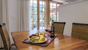 A dining table with a fruit platter, cheese, and wine glasses, set in a room with wicker chairs, a view of greenery outside, and a ceiling light.