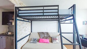 Una cama litera con colchón y almohadas blancos, pared con diseño de ladrillo y una zona de cocina con armarios y lavabo.