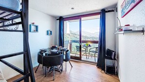 Una habitación con una cama litera, una mesa de comedor con sillas, vista a un paisaje montañoso a través de una puerta de cristal y un refrigerador en la pared.