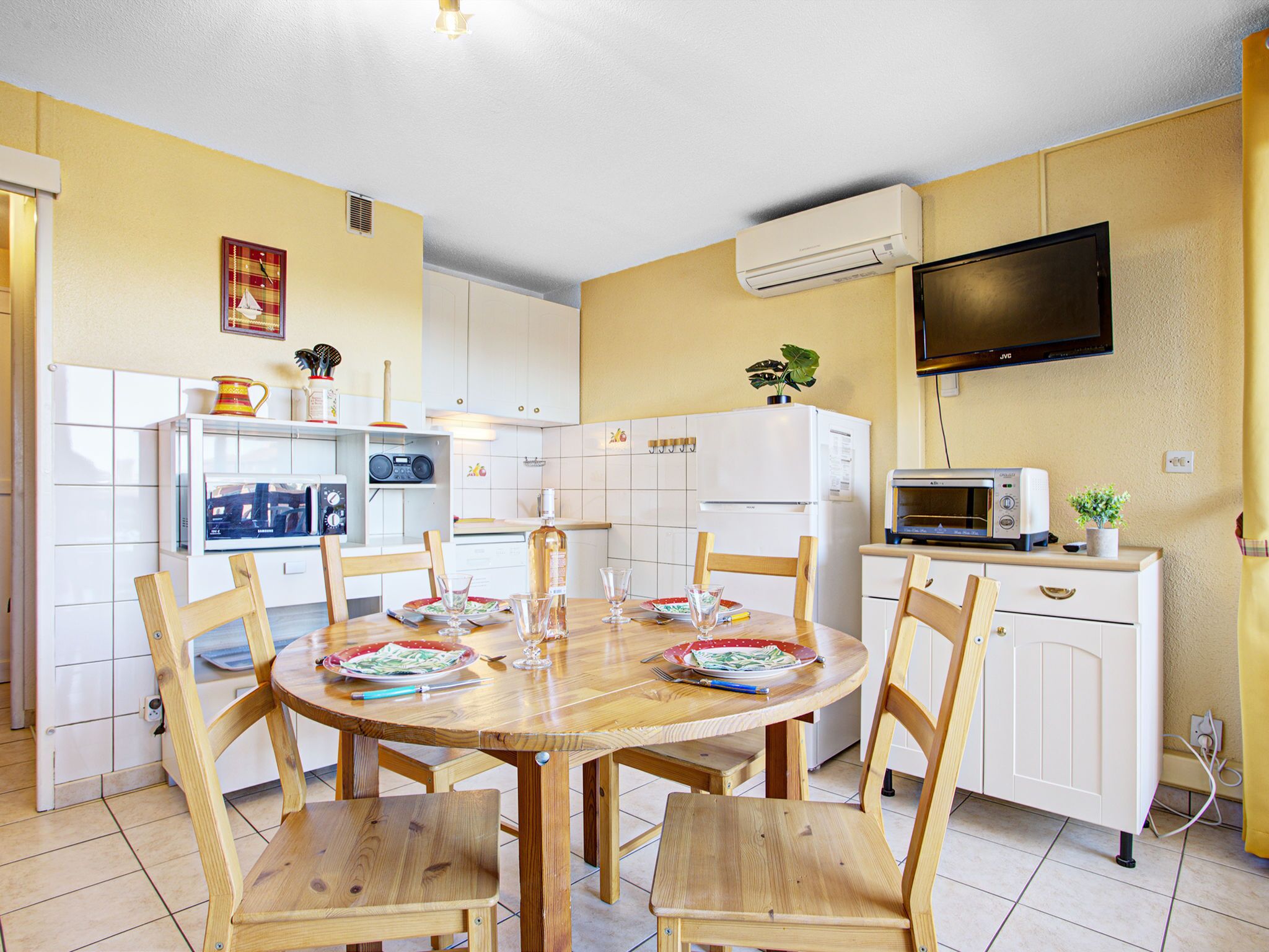 A compact kitchen with white cabinets, a dining table with chairs, a microwave, a toaster, and a flat-screen TV on the wall.