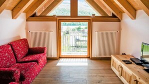 A room with a sloped wooden ceiling, a red floral sofa, a wooden table with a TV, and a view through a glass door to an outdoor area. - Albergo Diffuso - Cjasa de Barce by Interhome (Barcis Lake)