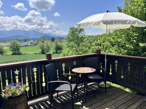 En träveranda med ett bord och två stolar, ett vitt parasoll och utsikt över ett grönskande landskap med träd och berg.