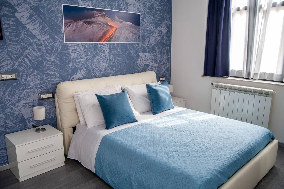 Un dormitorio con cama, mesita de noche, lámpara y un cuadro colgado en la pared.