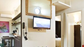 A modern hotel room with a flat-screen TV mounted on the wall, a desk with a telephone, and a small table with decorative items.
