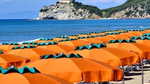 A beach with rows of orange umbrellas and lounge chairs, a rocky cliff with a building on top, and a clear blue sea.