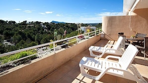 Un balcone con poltrone da relax bianche, un tavolino e vista su un paesaggio verde con alberi e montagne.