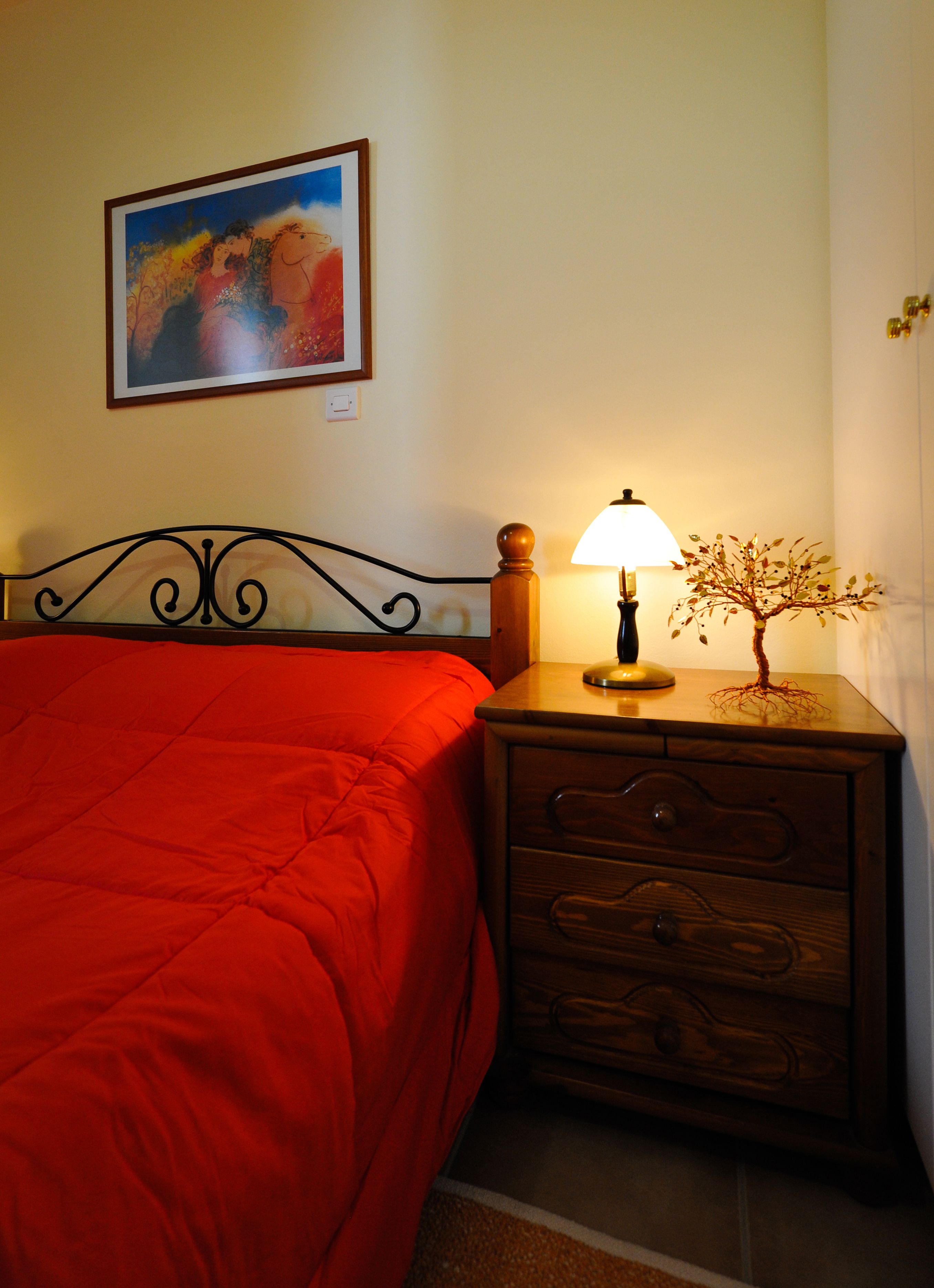 Un dormitorio con colcha roja, mesita de noche con lámpara, una planta pequeña y un cuadro en la pared.