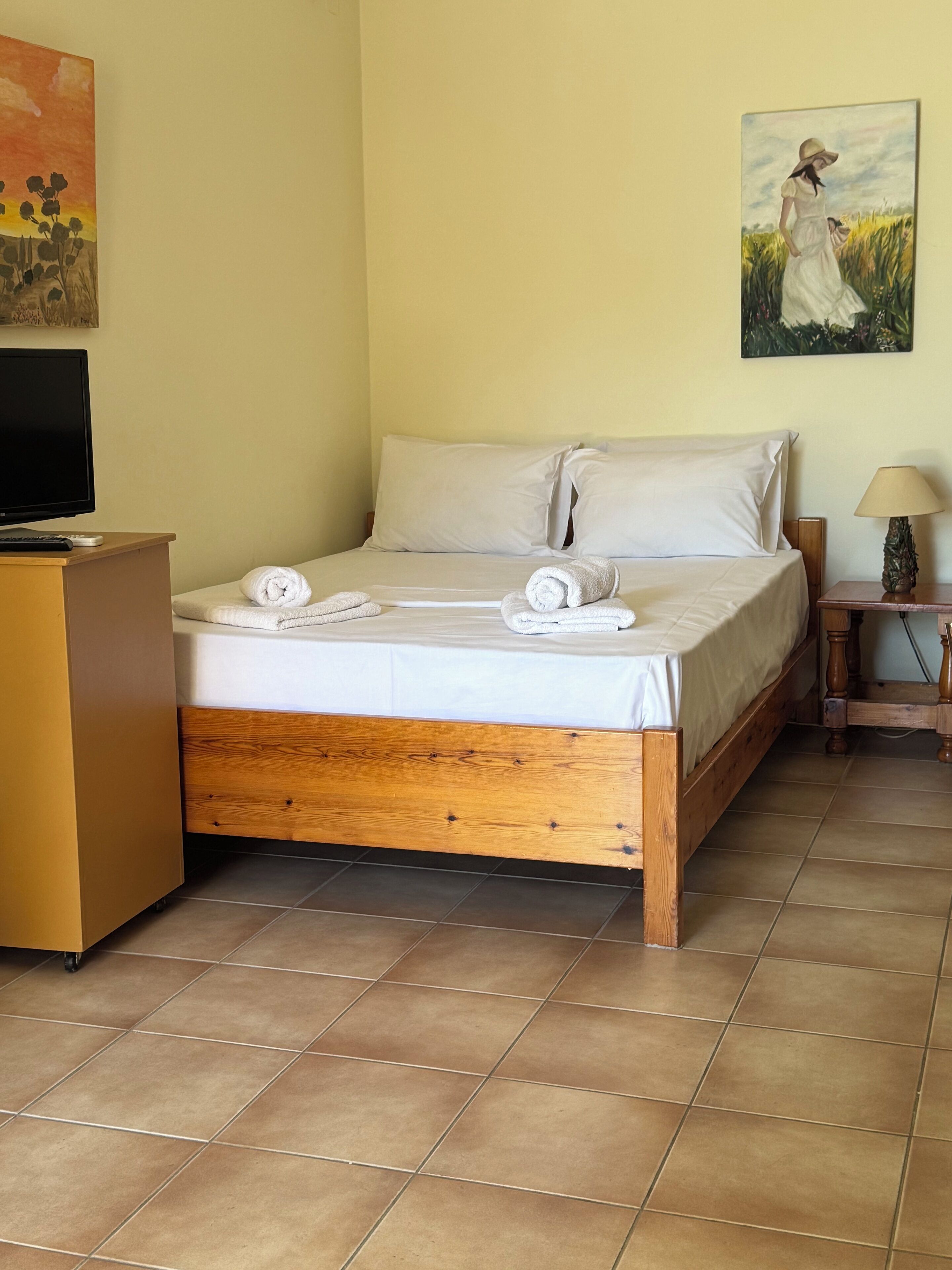 Una cama bien hecha con sábanas y toallas blancas, un cabecero de madera, una mesita de noche con una lámpara y un cuadro en la pared.