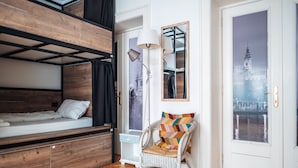 Una habitación con una cama litera, una silla con un cojín de colores, una lámpara de pie y vistas a un edificio a través de una puerta de vidrio.