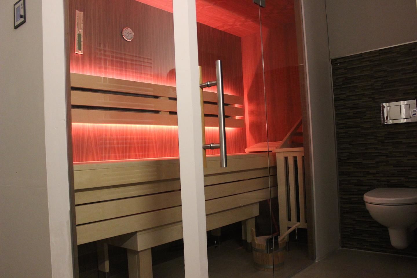 Een sauna met houten banken en een glazen scheiding, een houten bank met metalen frame, en een toilet met een donkere betegelde muur.