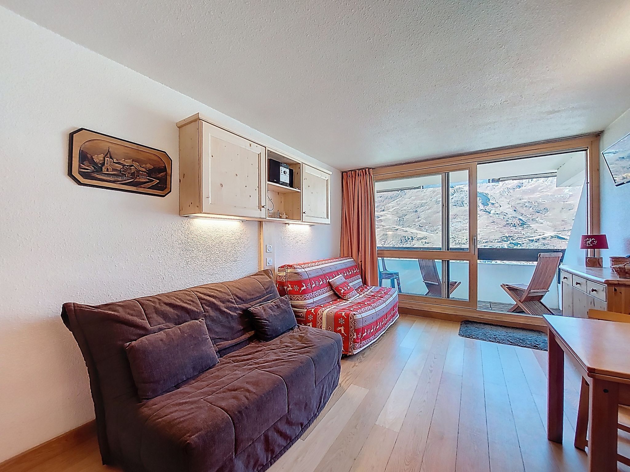 Uma sala de estar aconchegante com um sofá marrom, um banquinho estampado, armários de madeira e uma janela grande com vista para uma paisagem nevada.