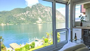 Una stanza con una grande finestra che si affaccia su un lago e montagne. C'è un divano bianco con cuscini, un tavolino con una bottiglia e bicchieri, e uno specchio sopra il tavolino.