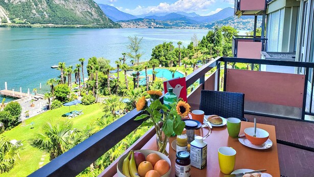 Un balcone con tavolo apparecchiato per la colazione, vista su un lago, circondato da vegetazione e montagne.