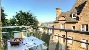 Un balcon avec une table dressée pour deux personnes, un panier rempli de victuailles et une bouteille d’eau. Le balcon donne sur un bâtiment en pierre, surmonté d’un toit en tuiles.