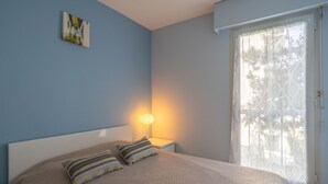 Een slaapkamer met een bed, kussens, een bedlampje en een raam met een gordijn met bladmotief.