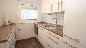 Et lille køkken med hvide skabe, en træbordplade, en stålvask og et vindue med gardin.