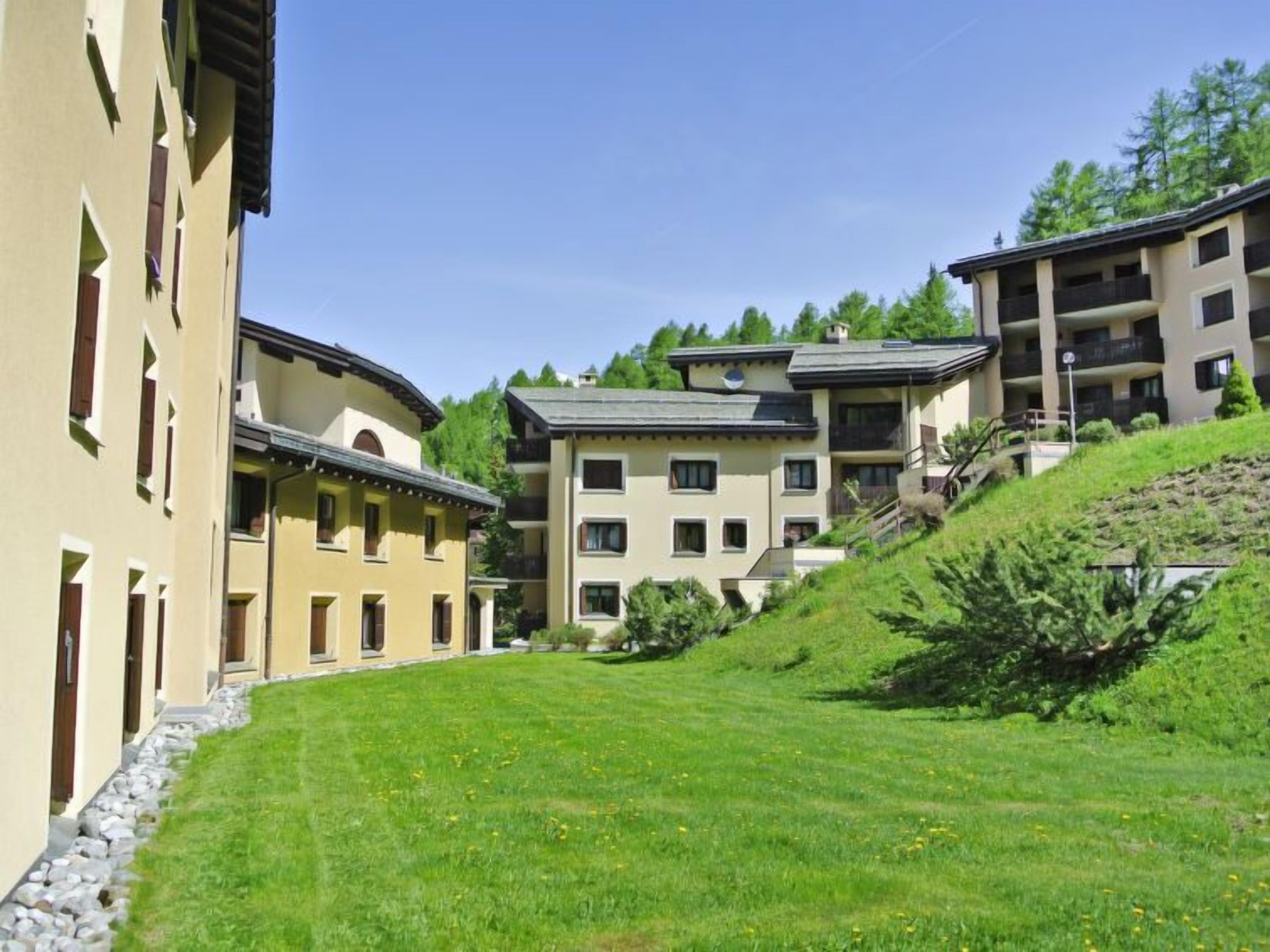 A row of multi-story buildings with balconies, a grassy courtyard, and surrounding trees.