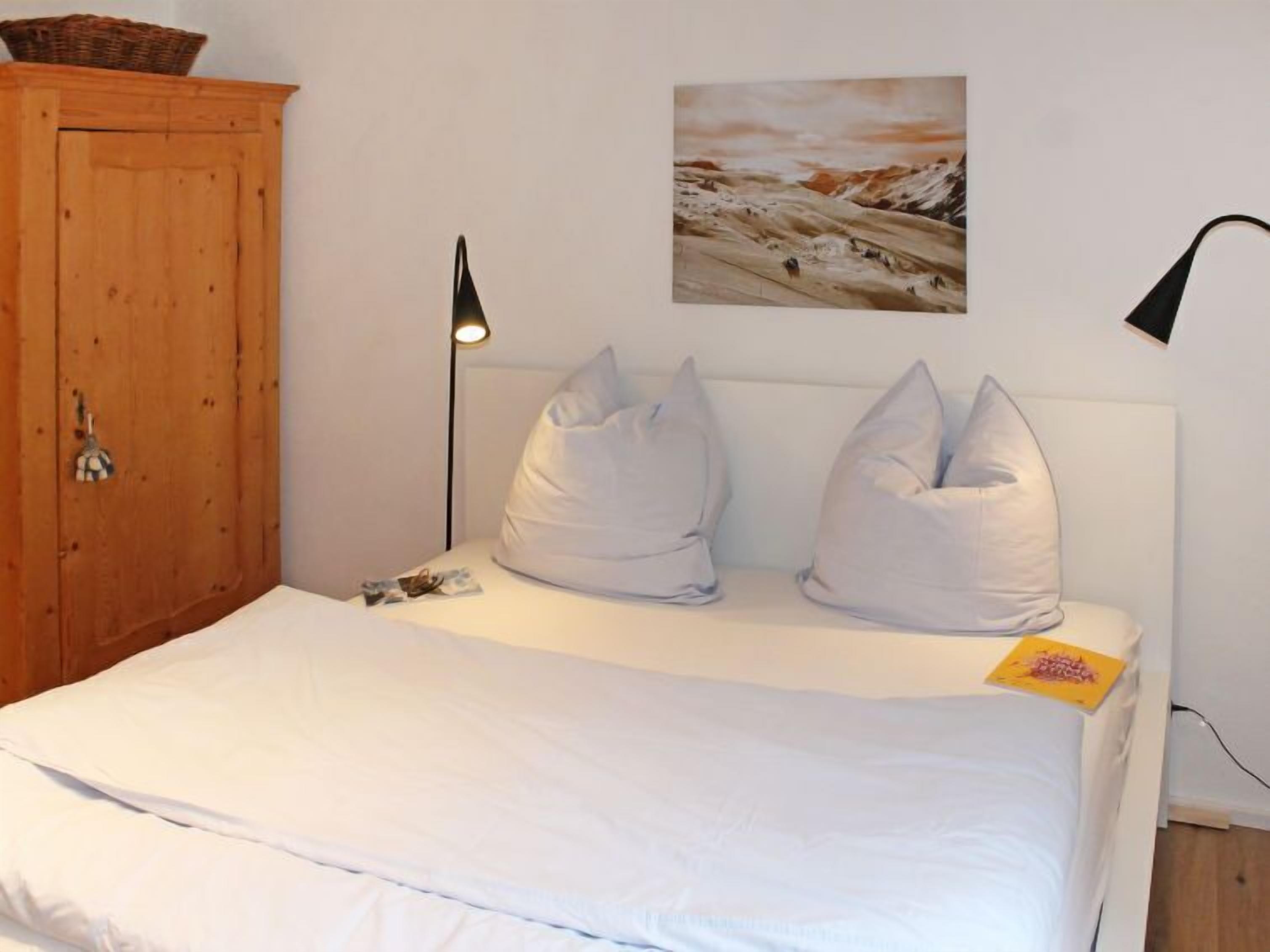 Une chambre avec une armoire en bois, un lit avec une literie blanche, deux oreillers, une lampe de chevet et un tableau au mur.