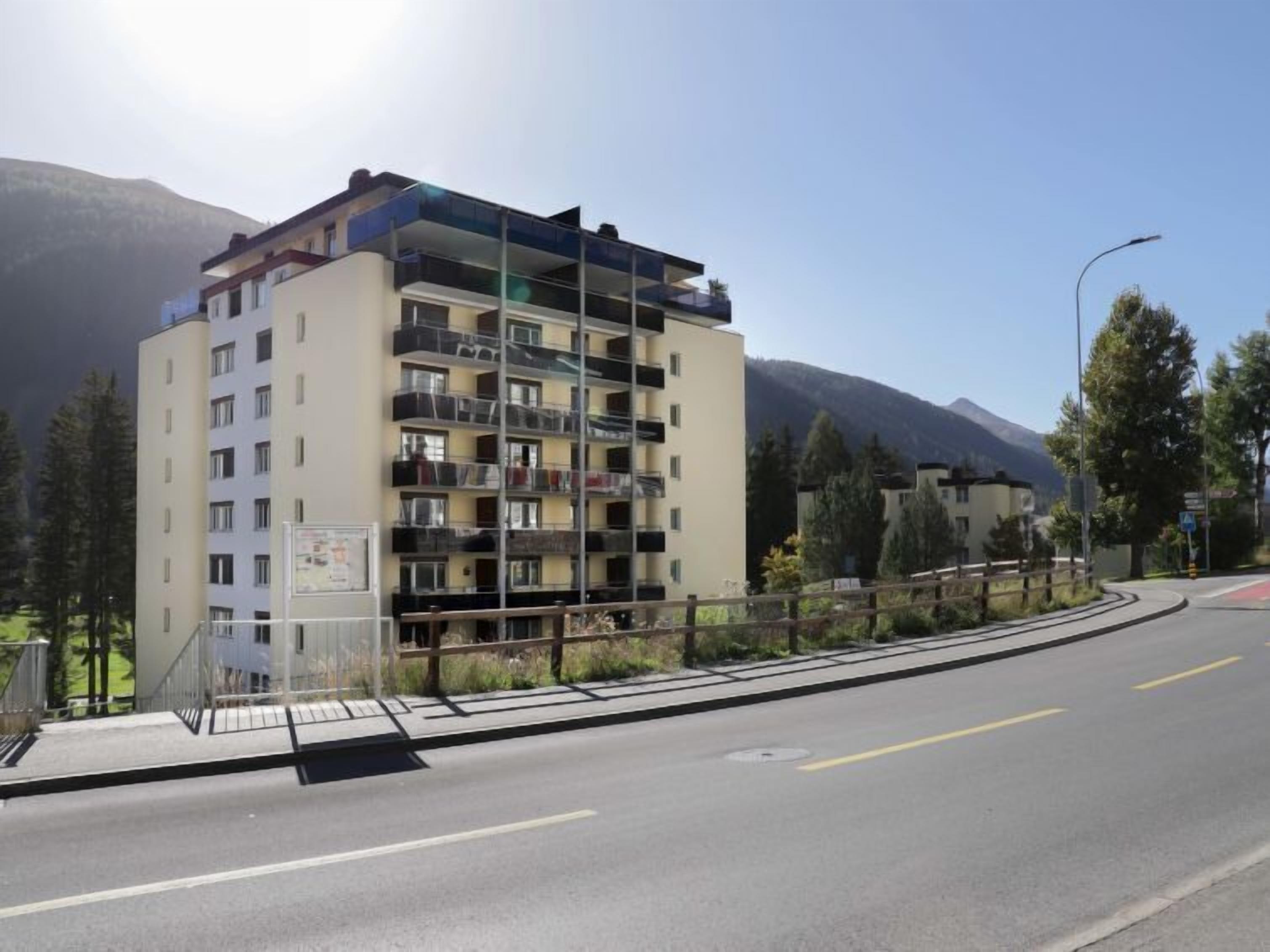 Un edificio de varios pisos con balcones, una calle en primer plano y montañas al fondo.