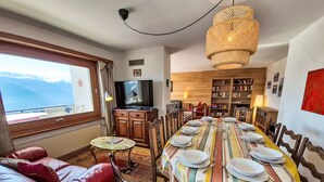 A dining area with a long table set for six, a television, a bookshelf, and a view of mountains through a window. - Chalet Aglaia by Interhome (Crans-Montana)