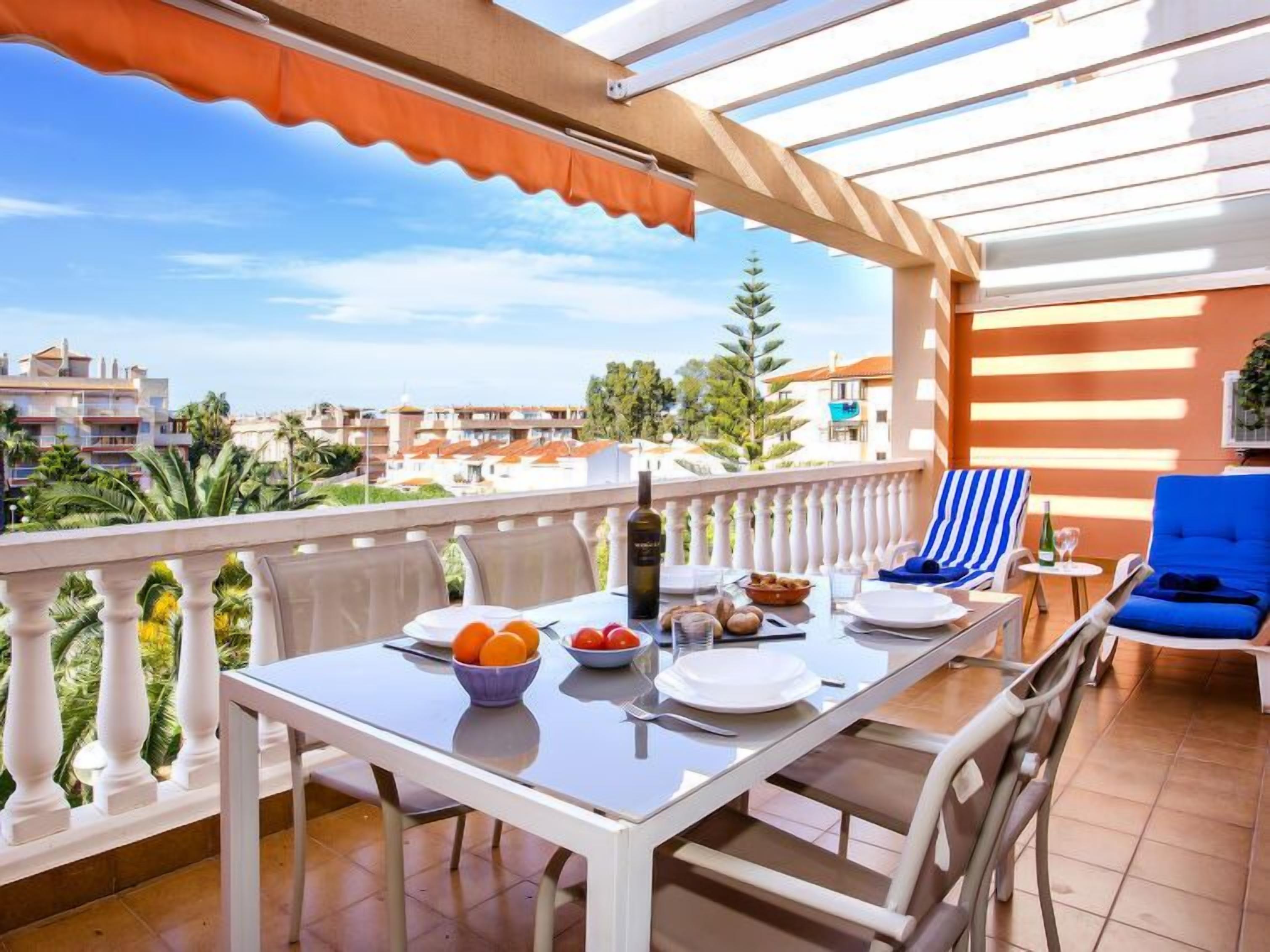Un balcon avec une table préparée pour le petit-déjeuner, comprenant des oranges et des tomates, ainsi qu’une bouteille d’huile d’olive.